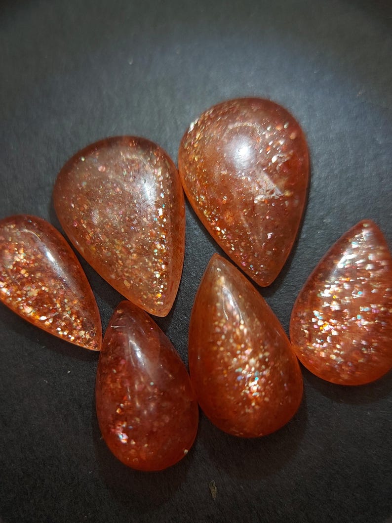 May include: Six teardrop-shaped sunstone cabochons with a shimmering, iridescent finish. The stones are a mix of orange and red hues.