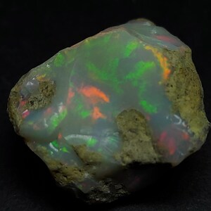 Op de afbeelding: Een ruwe opaal edelsteen met een ruw, getextureerd oppervlak. De opaal toont levendige interne kleuren, waaronder groen, oranje en rood, tegen een melkachtige witte basis. De steen heeft een onregelmatige vorm, die zijn natuurlijke vorm benadrukt.