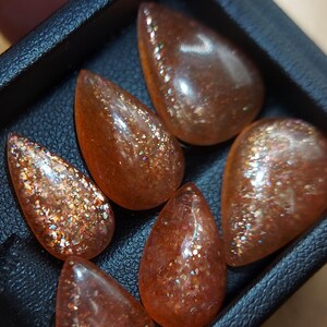 May include: Six teardrop-shaped sunstone cabochons in a black leather box. The stones are a translucent orange color with a glittery, iridescent finish.