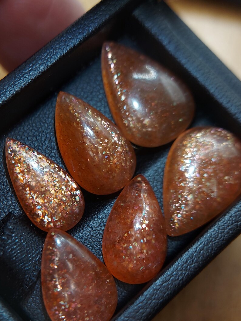 May include: Six teardrop-shaped sunstone cabochons in a black leather box. The stones are a translucent orange color with a glittery, iridescent finish.
