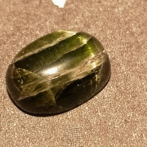 May include: A dark green oval-shaped gemstone with a striated pattern. The gemstone is polished and has a glossy finish.