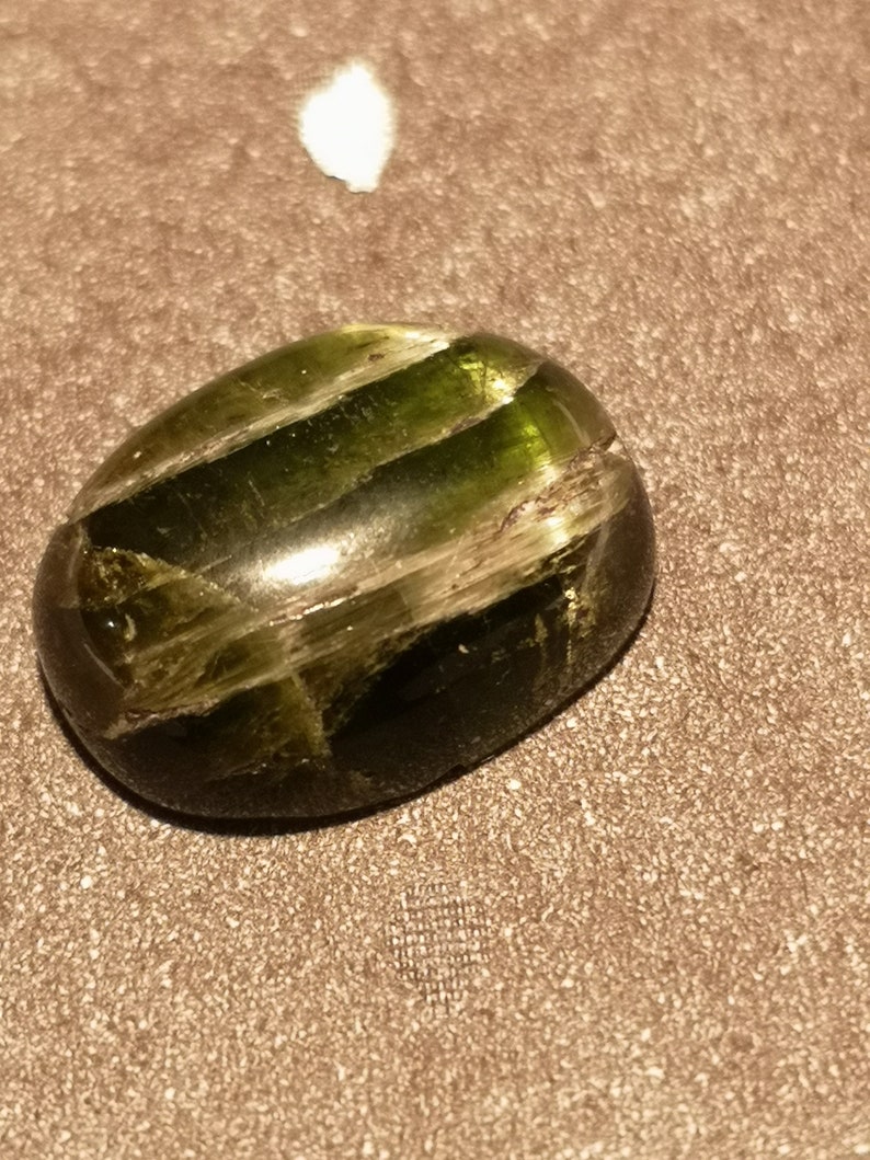 May include: A dark green oval-shaped gemstone with a striated pattern. The gemstone is polished and has a glossy finish.