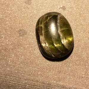 May include: A green gemstone cabochon with a striated pattern. The stone is oval shaped and has a smooth, polished surface.