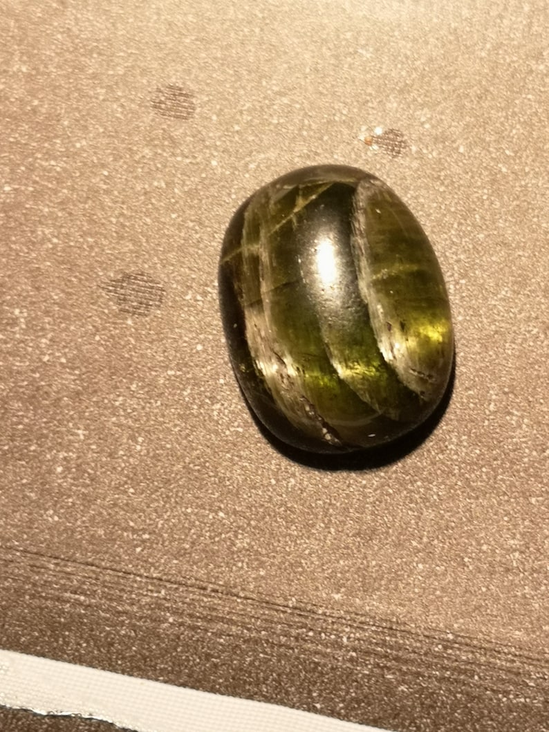 May include: A green gemstone cabochon with a striated pattern. The stone is oval shaped and has a smooth, polished surface.