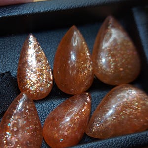 May include: Six teardrop-shaped sunstone gemstones with a shimmering, iridescent finish. The stones are a mix of orange, brown, and gold colors.