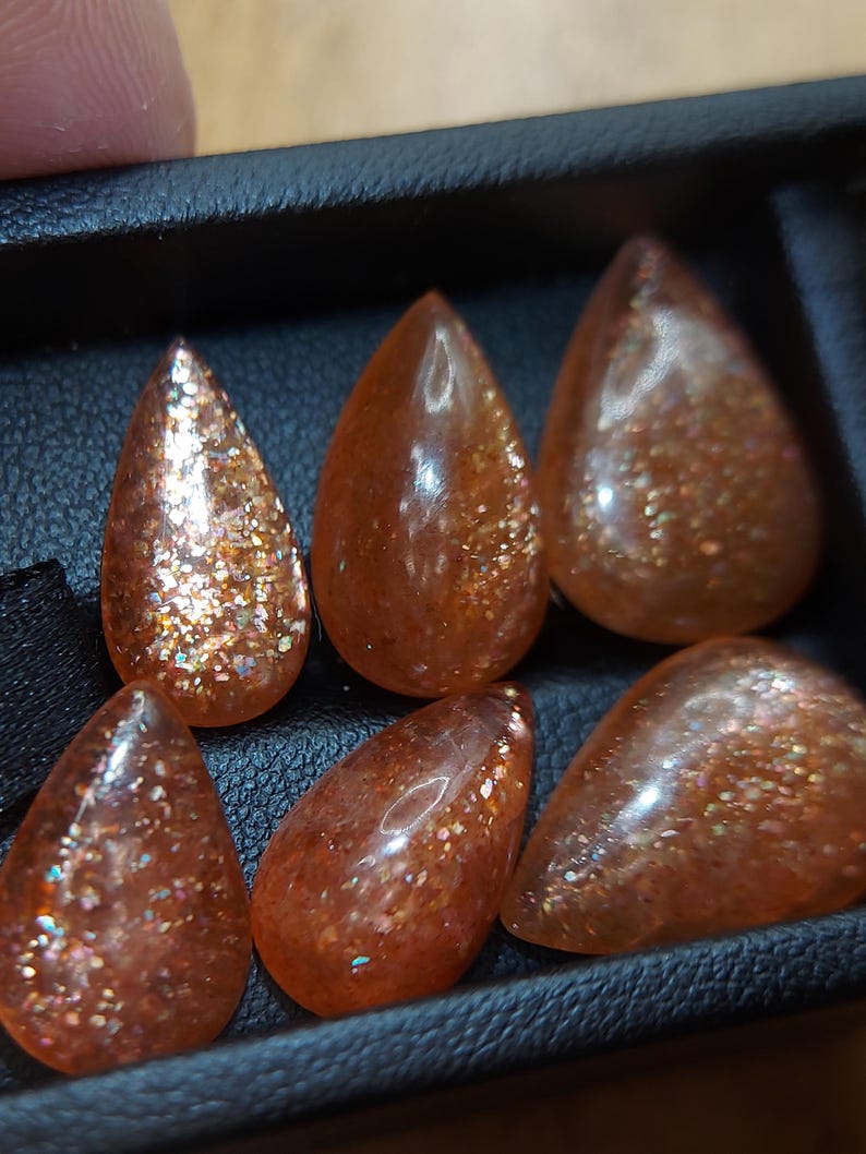 May include: Six teardrop-shaped sunstone gemstones with a shimmering, iridescent finish. The stones are a mix of orange, brown, and gold colors.