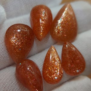 May include: Six teardrop-shaped sunstone gemstones with a shimmering, iridescent finish. The stones are a mix of orange and brown with a glittery appearance.