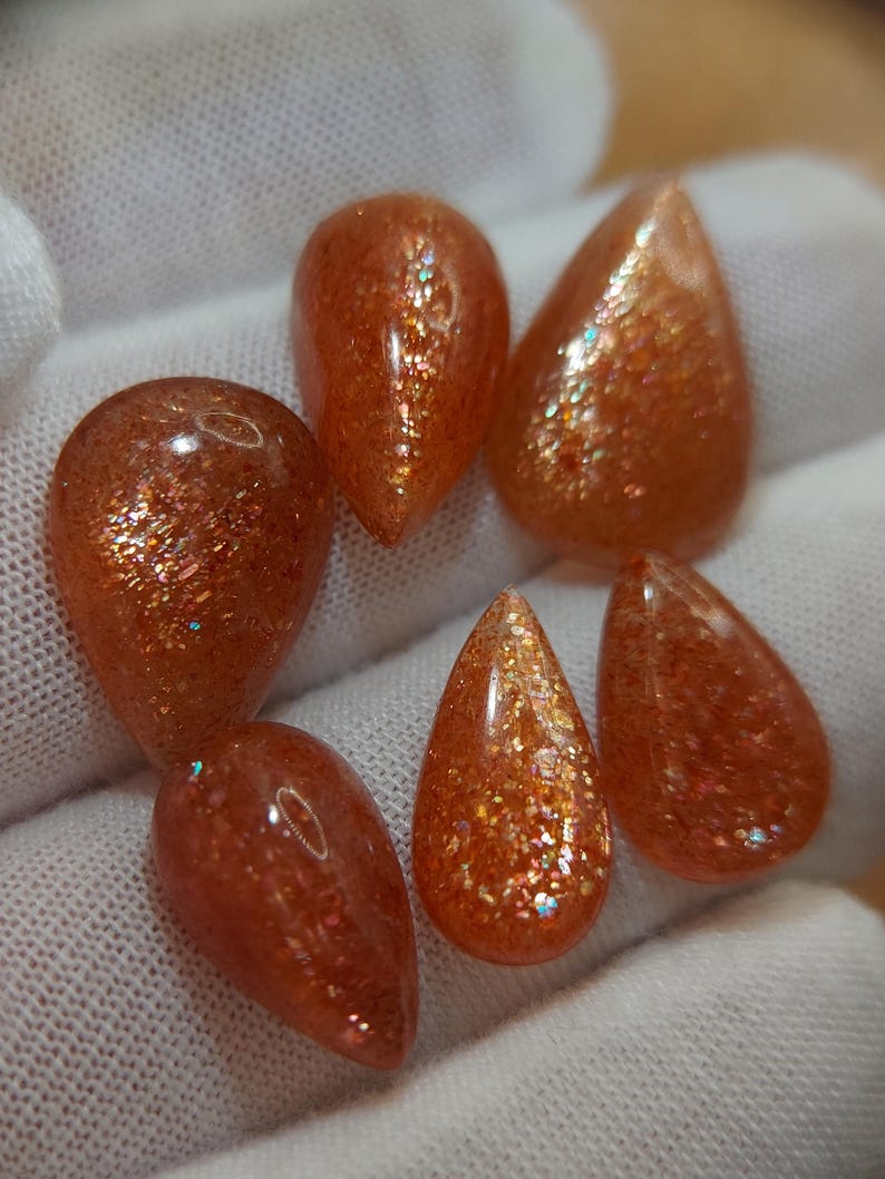 May include: Six teardrop-shaped sunstone gemstones with a shimmering, iridescent finish. The stones are a mix of orange and brown with a glittery appearance.