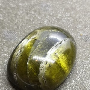 May include: A green and black gemstone with a smooth, oval shape. The stone has a striated pattern and a glossy finish.