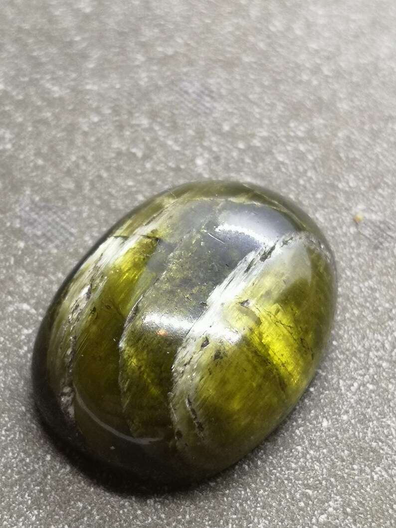May include: A green and black gemstone with a smooth, oval shape. The stone has a striated pattern and a glossy finish.