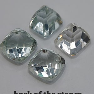 May include: Four clear, faceted, square-shaped gemstones with a light blue hue. The back of the stones are shown in the image.