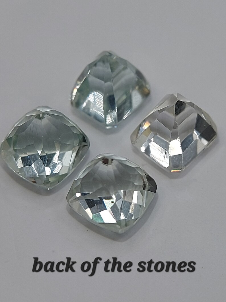 May include: Four clear, faceted, square-shaped gemstones with a light blue hue. The back of the stones are shown in the image.