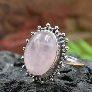 May include: A silver ring featuring a large, oval rose quartz gemstone. The light pink stone is set in a detailed, ornate silver setting with a beaded border. The ring is displayed on a dark, textured surface.
