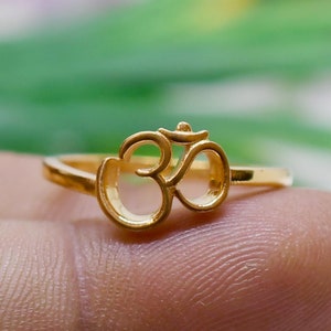 Peut inclure: Une bague dorée ornée du symbole Om. La bague a un anneau fin, et le symbole Om est le point central, avec son design complexe clairement visible. L'arrière-plan est doux et flou.