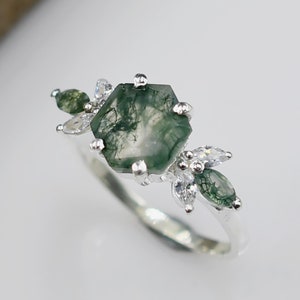 May include: A silver ring with a green moss agate gemstone and small clear cubic zirconia stones. The ring has a delicate floral design.
