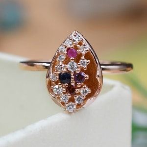 Può includere: Anello in oro rosa con castone a forma di goccia. Il castone è riempito con un gruppo di piccole pietre preziose colorate, tra cui pietre bianche, rosse, viola e blu scuro. L'anello è posto su uno sfondo bianco.