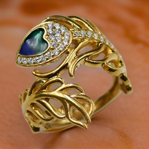 May include: A gold ring featuring a heart-shaped blue and green stone set within a halo of clear cubic zirconia stones. The ring is crafted with a delicate, intricate feather design.