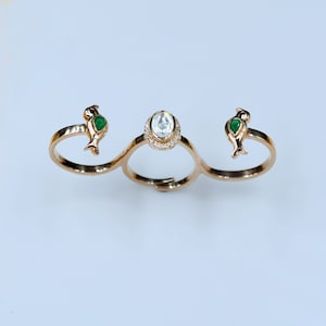 May include: A gold-toned, three-finger ring featuring a central oval-shaped, clear gemstone surrounded by small, clear stones. Two bird-shaped accents with green and red details flank the central stone. The ring is set against a white background.