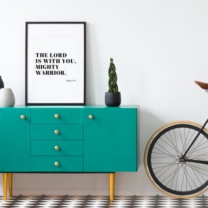 Pu&ograve; includere: Una stampa incorniciata nera con il testo "The Lord is with you, mighty warrior. Judges 6:12" appesa a un muro bianco sopra una cassettiera blu turchese con pomelli dorati. Nell'immagine sono presenti anche una pianta in vaso e una bicicletta.