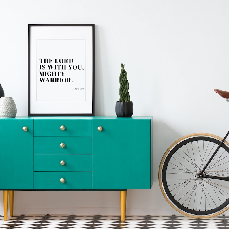 Pu&ograve; includere: Una stampa incorniciata nera con il testo "The Lord is with you, mighty warrior. Judges 6:12" appesa a un muro bianco sopra una cassettiera blu turchese con pomelli dorati. Nell'immagine sono presenti anche una pianta in vaso e una bicicletta.