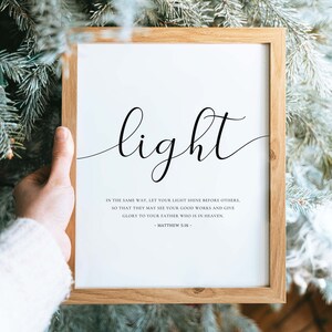 Może przedstawiać: Biały wydruk w ramie z napisem "light" w ozdobnym, pochylonym piśmie. Wydruk zawiera werset biblijny z Ewangelii Mateusza 5:16: "Tak niech świeci wasze światło przed ludźmi, aby widzieli wasze dobre uczynki i chwalili Ojca waszego, kt&oacute;ry jest w niebie."