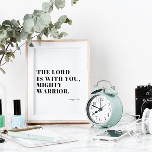 Pu&ograve; includere: Una stampa incorniciata con il testo "The Lord is with you, Mighty Warrior. Judges 6:12" in testo nero su sfondo bianco. Una sveglia blu chiaro con lancette e numeri neri &egrave; in primo piano.