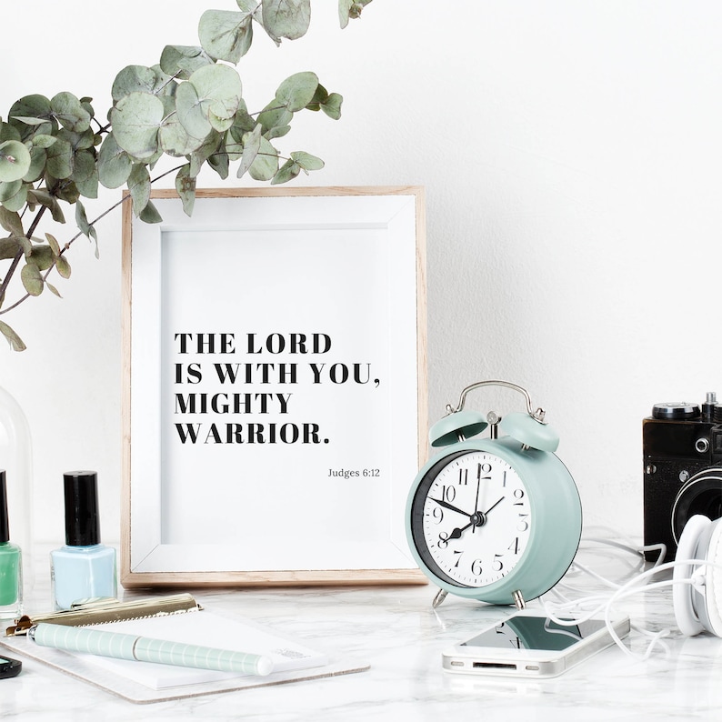 Pu&ograve; includere: Una stampa incorniciata con il testo "The Lord is with you, Mighty Warrior. Judges 6:12" in testo nero su sfondo bianco. Una sveglia blu chiaro con lancette e numeri neri &egrave; in primo piano.