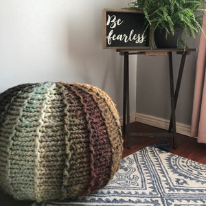 May include: A large, round, hand-knitted pouf in shades of brown, green, and blue sits on a patterned rug. A small wooden sign with the words "Be fearless" is displayed near a potted fern. The pouf is a cozy home decor item.