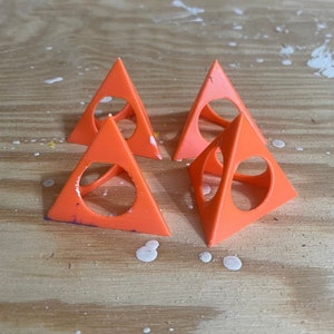 May include: Four orange plastic geometric shapes in the form of a pyramid with a hole in the center of each face. The pyramids are sitting on a wooden surface.