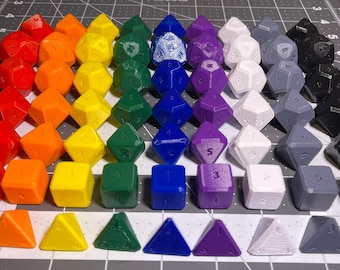May include: A collection of multi-sided gaming dice arranged in a spectrum of colors including red, orange, yellow, green, blue, purple, white, gray, and black. The dice feature various geometric shapes and numerical markings on their surfaces.