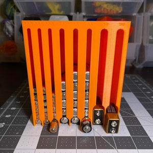 May include: An orange 3D-printed battery organizer holding various battery sizes, including AA, AAA, C, and 9-volt batteries. The organizer is designed with vertical slots to separate and store batteries neatly. The organizer is on a black and gray checkered surface.