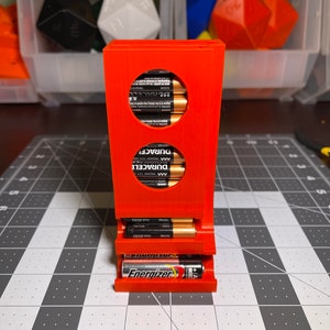 May include: An orange plastic battery organizer with three compartments. The top two compartments are circular and hold AA batteries. The bottom compartment is rectangular and holds AAA batteries. The batteries are labeled "Duracell" and "Energizer".