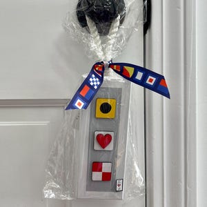 May include: A decorative door knob cover with a nautical theme. The cover is wrapped in clear plastic and tied with a ribbon featuring colorful geometric patterns. The cover has three shapes: a yellow square with a black circle, a red heart, and a red and white checkered square.