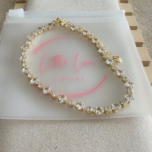 May include: A gold-tone tennis bracelet featuring a continuous row of clear, square-cut cubic zirconia stones. The bracelet includes a small heart-shaped charm near the clasp and is presented on a translucent pouch with the text Little Lou JEWELRY.