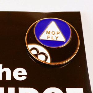 May include: A black and gold enamel pin featuring a black 8-ball with a blue triangle on top. The triangle has the words "Mop Fly" written in white. The pin is on a black background with the words "The Midge Pin" in white.