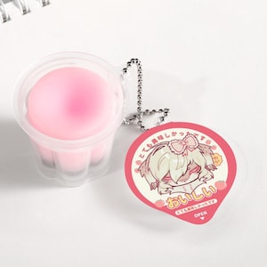 May include: A pink, jelly-like dessert in a clear plastic cup with a decorative tag. The tag features a cartoon character and Japanese text. A silver chain is attached to the cup and tag.