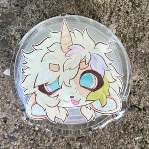 May include: A circular clear acrylic phone grip featuring a printed illustration of a white unicorn character with shaggy hair, a single horn, large blue eyes, and a small pink tongue sticking out, finished with a subtle glittery overlay.