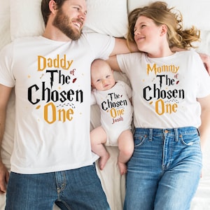 May include: A family of three wearing white t-shirts with a Harry Potter themed design. The shirts say "Daddy of the Chosen One", "Mommy of the Chosen One", and "The Chosen One Jonah".