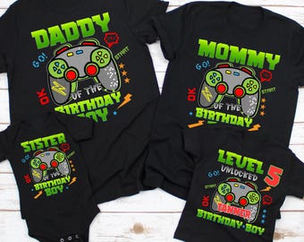 May include: A set of four black t-shirts with a video game theme. The shirts say "Daddy of the Birthday Boy", "Mommy of the Birthday Boy", "Sister of the Birthday Boy", and "Level 5 Unlocked Tanner Birthday Boy".
