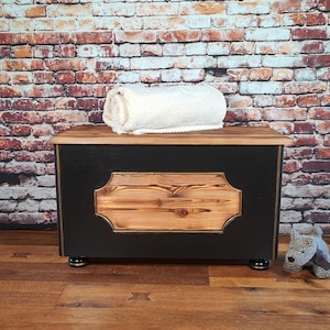 May include: Black wooden storage chest with a gold trim and a natural wood panel on the front. The chest is sitting on a wooden floor and has a white towel on top.