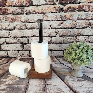 May include: A wooden toilet paper holder with a black metal rod and a gold accent. The holder is designed to hold three rolls of white toilet paper. One roll is lying on the wooden surface. The background features a brick wall and a small plant.
