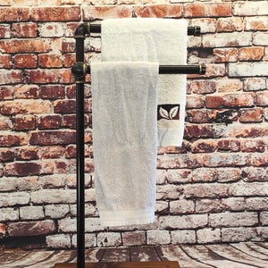 Pode incluir: Um toalheiro de metal preto e madeira com duas toalhas brancas. O suporte tem uma base retangular de madeira e uma estrutura semelhante a um cano. As toalhas estão penduradas nas barras horizontais.