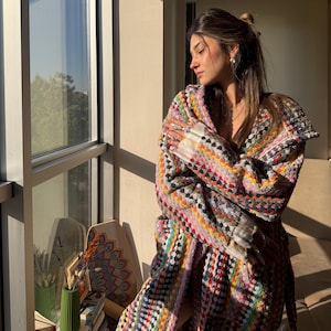 May include: A vibrant, patterned dressing gown with a hood. The gown displays a mix of bright colours, including pink, yellow, blue, and green. The gown is worn indoors near a large window, with sunlight illuminating the scene.