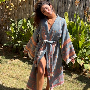 May include: A blue kimono-style robe featuring a woven pattern of orange and cream-colored celestial motifs, including stars and moons. The garment has wide sleeves, a matching tie belt at the waist, and white fringe trim along the hem and cuffs.