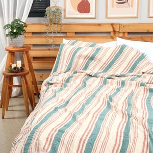 Puede incluir: Una manta color crema con rayas verticales en verde azulado y rojo, sobre una cama. La manta tiene un borde con flecos. La cama está contra un cabecero de madera, con arte enmarcado en la pared. Un pequeño taburete de madera con una planta y velas está en primer plano.