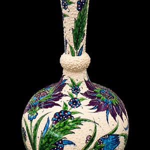 Op de afbeelding: Een keramische vaas met een crèmekleurige basis en een textuur met kleine stippen. De vaas is voorzien van handgeschilderde bloemmotieven in tinten dieppaars, levendig groen en blauw, met lange, slanke bladeren en kleine, ronde bloesems.