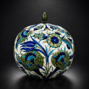 May include: A round ceramic jar with a matching lid featuring a pointed finial. The surface is decorated with a hand-painted floral pattern in shades of deep blue, turquoise, and olive green against a cream-colored background.