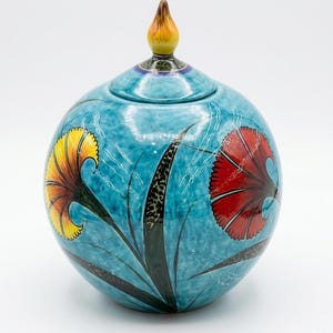 Pode incluir: Um vaso de cerâmica turquesa redondo com tampa, com desenhos florais pintados à mão. O vaso tem uma flor amarela e vermelha e um remate castanho e amarelo. Uma peça decorativa.