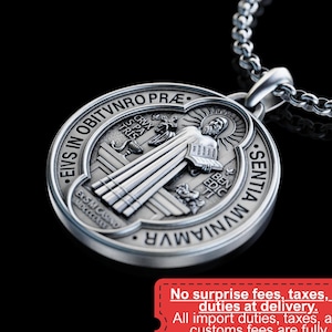 May include: A silver pendant necklace with a circular medallion displaying religious iconography and Latin text. The pendant is suspended from a chain. A red tag with text about import duties is also visible.