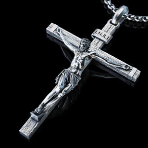 Puede incluir: Un colgante de crucifijo en tono plateado que presenta una figura detallada de Jesús sobre una cruz texturizada con las letras I.N.R.I. inscritas en una pequeña placa sobre la cabeza, suspendido de un collar de cadena de metal plateado.